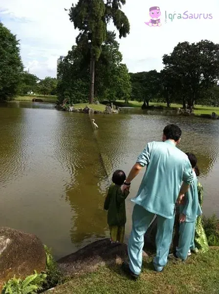 Daddy in Hari Raya clothes holding a toddler's hand at the edge of a lake, with an egret on the water
