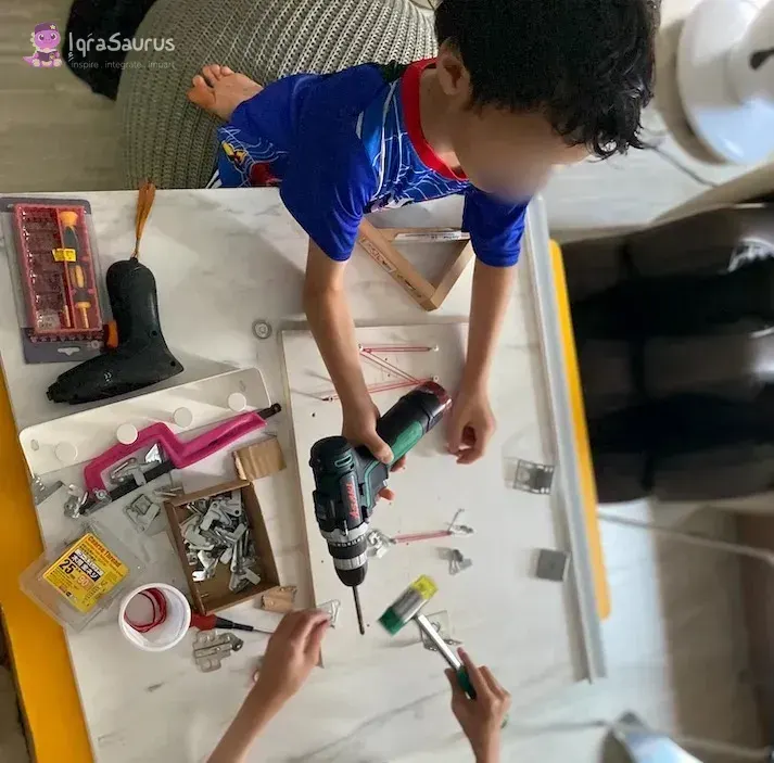 Qoyis, as a preschooler, using a cordless drill on a board, with hinges, brackets and screwdrivers on the table
