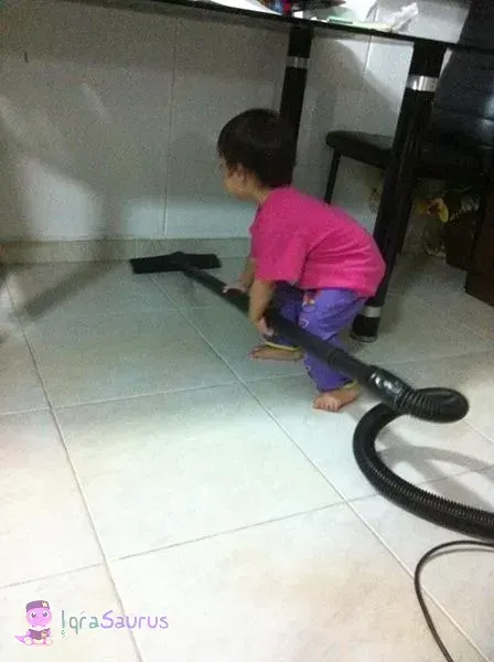 A toddler in a pink top pushes a vacuum cleaner hose under the dining table