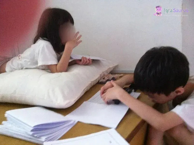 Two older children assembling paper booklets, one lying on a pillow and one bent over the pages