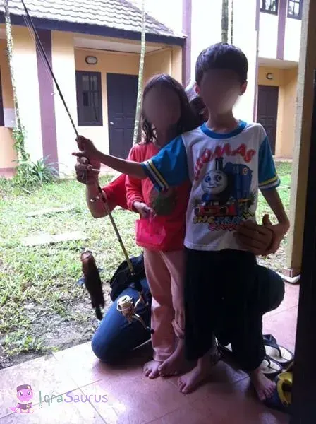 A boy holds up a fishing rod with a small catch, his sister standing beside him