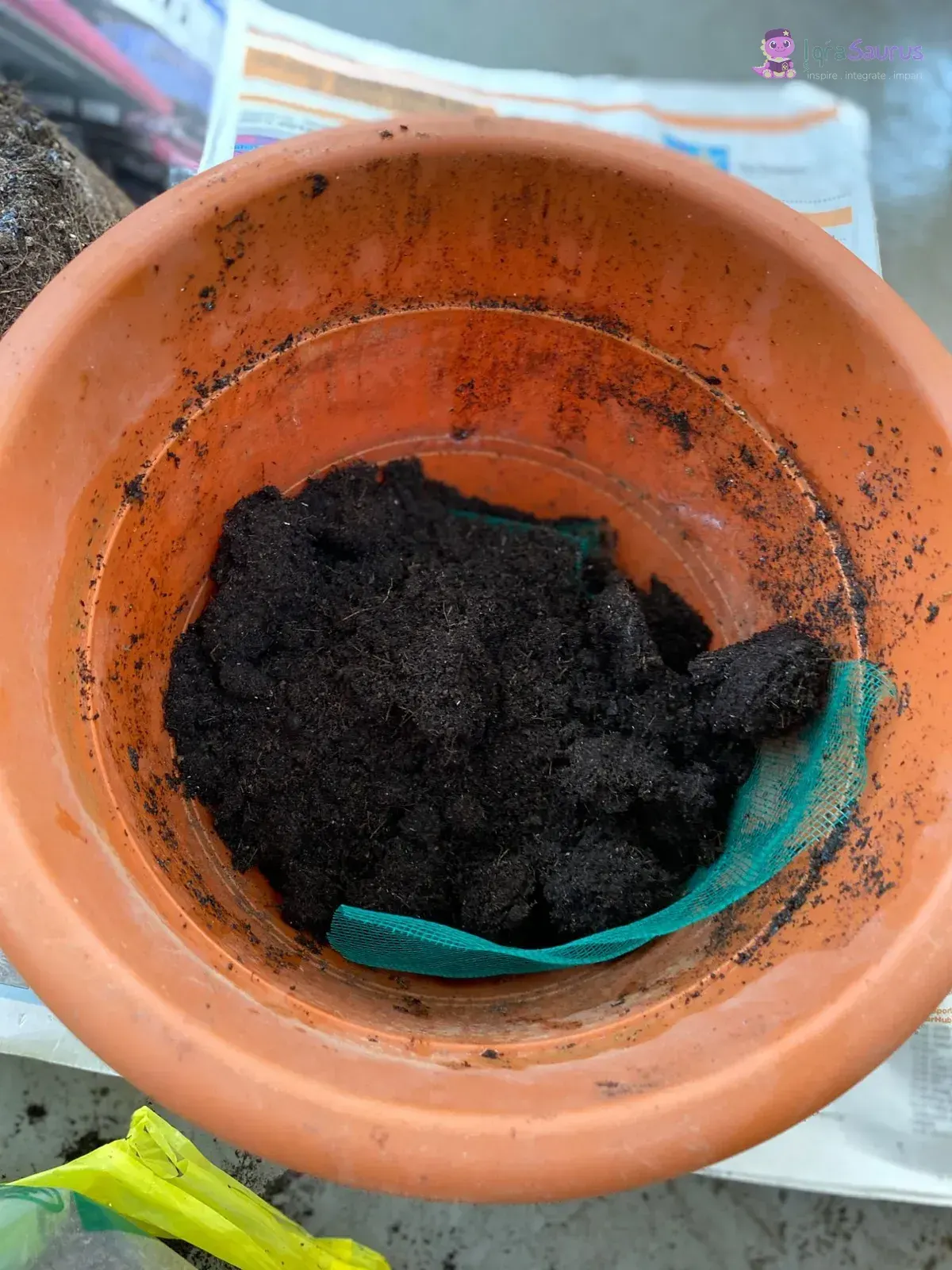 A clay pot lined with green net and half filled with dark potting soil